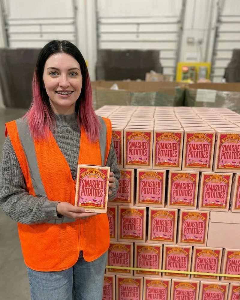 A volunteer holding a Teton Tater carton beside a pallet of cartons in a food bank warehouse